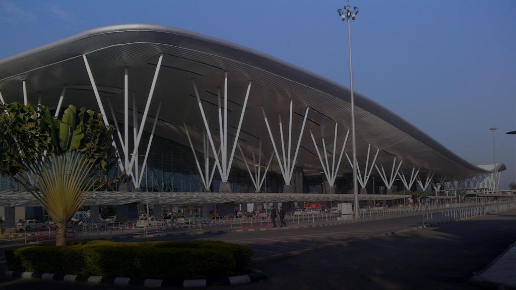 Airport terminal with a curved roof supported by branching tubular steel columns under a blue sky.