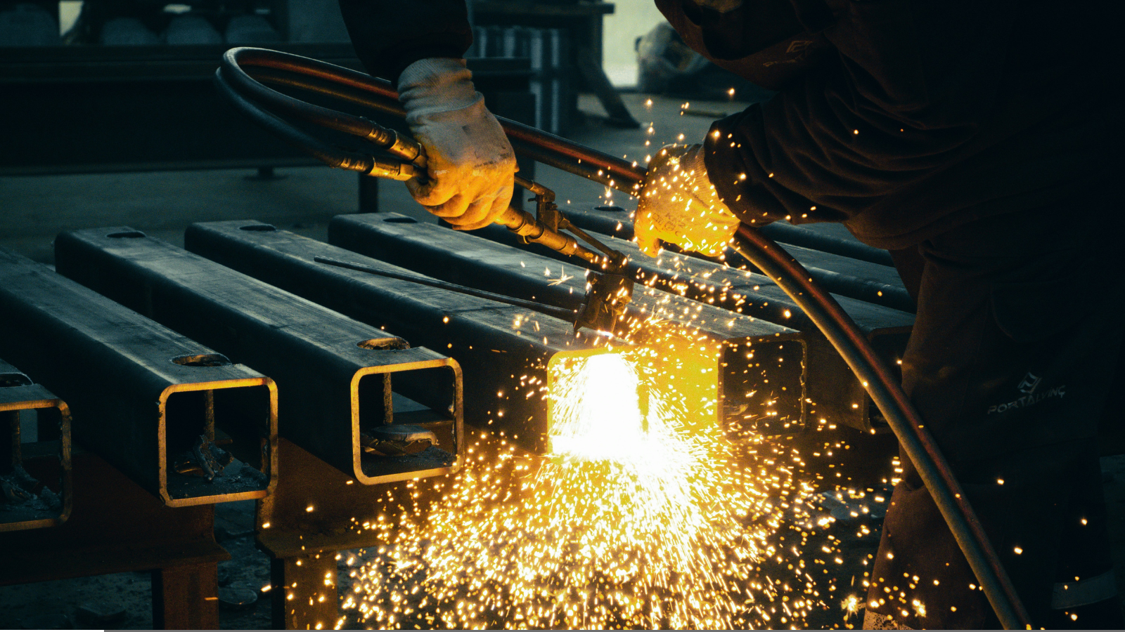 Home 7 Worker using a cutting torch on square steel tubes with a cascade of bright sparks in a workshop.