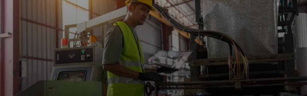 Worker in hard hat and safety vest inspecting equipment with a tablet inside a steel pipe factory.