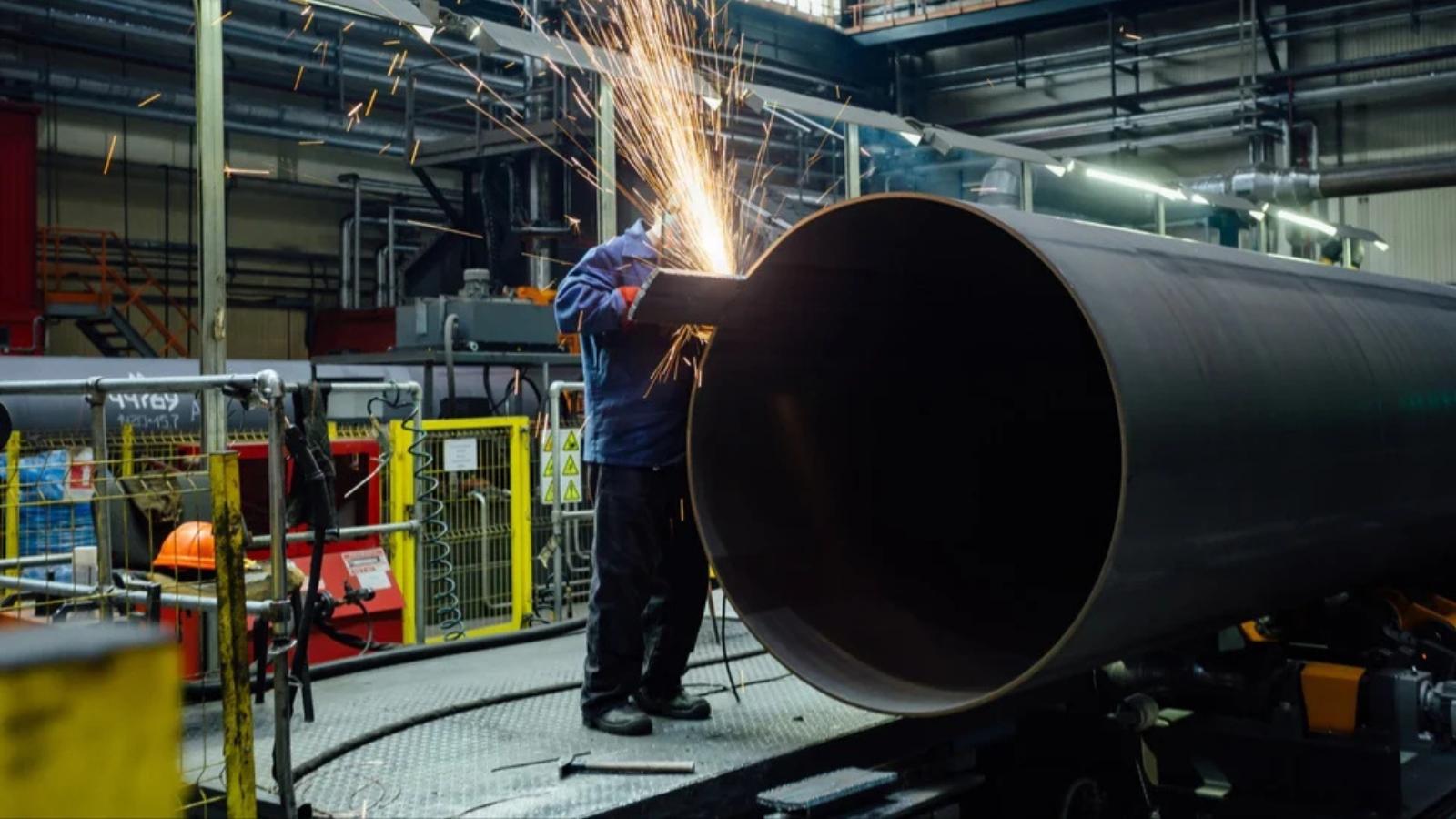 Worker welding a large steel pipe with bright sparks inside a pipe manufacturing factory.