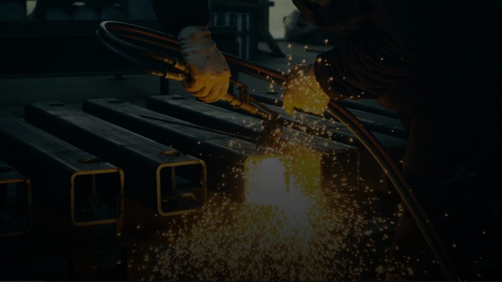 Worker torch-cutting square hollow steel sections with golden sparks in a dim workshop.