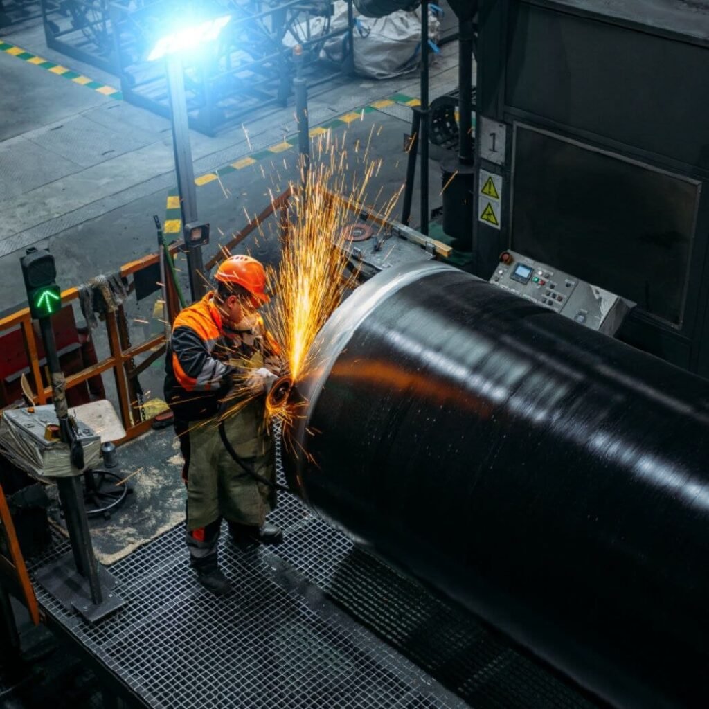 Worker in orange safety gear grinding the end of a large steel pipe with sparks in a factory.